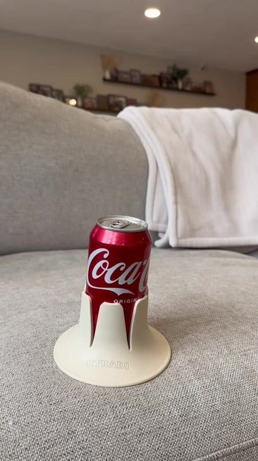 A full can standing in a bone-coloured Steadi on a couch cushion, a folded blanket over the sofa back behind it.